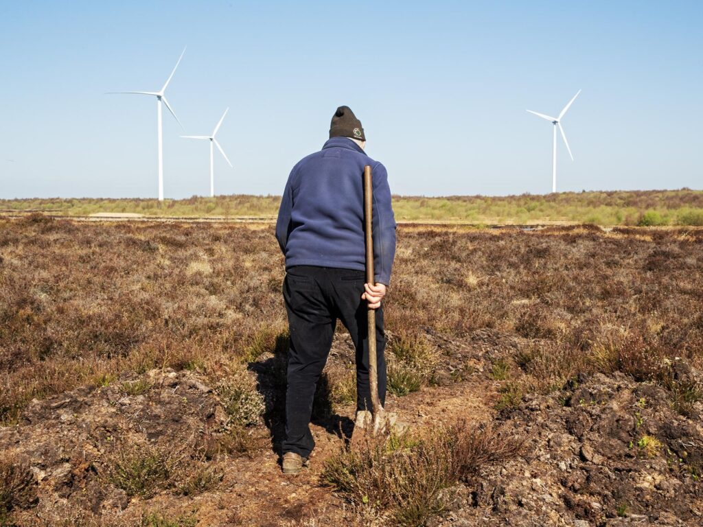 Michael O’Rourke, Walsh Island, photo Shane Hynan A man named Michael O’Rourke, is walking with a shovel on the bog Walsh Island with wind mills on ablue skyline