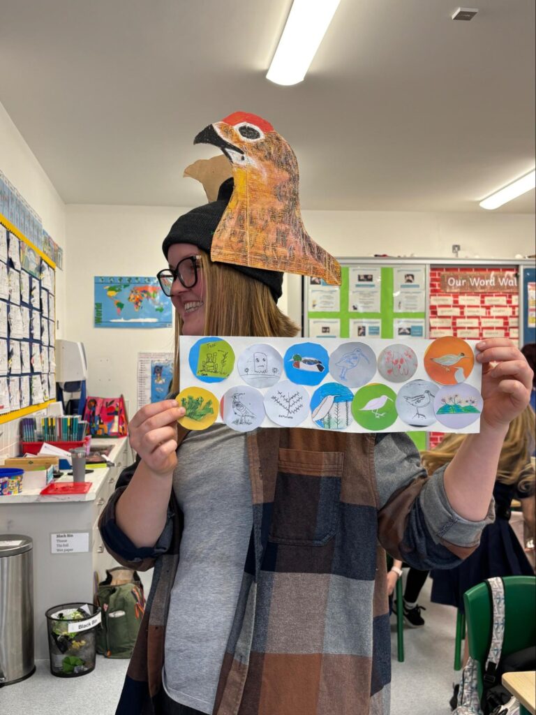Photo of Katie Smirnova wearing her Red Grouse Hat and showing the children’s artwork