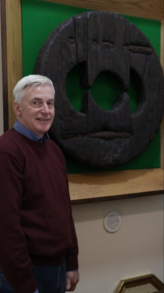 Photograph of Sean Mooney in front of the replica wheel, Kilteevan Community Centre.