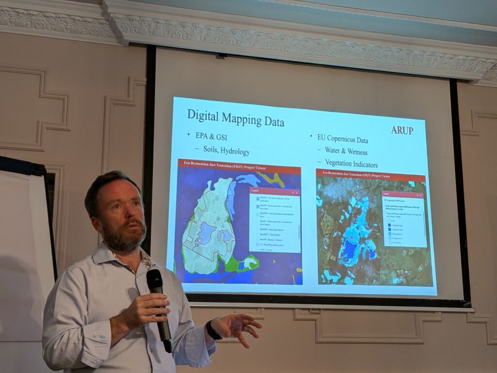 Man in blue shirt standing in front of a presentation slide that contains bullet points about digital mapping data including EPA & GSI maps on soils and hydrology as well as EU Copernicus Data maps that contain  water & wetness as well as vegetation index.