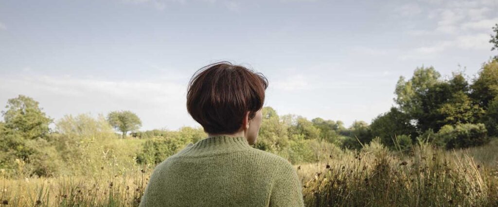 Woman wearing green cardigan with brown hair standing alone looking over long meadow grass and a landscape of trees and grasses.