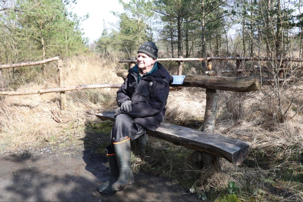 Hugh Shephard dressed in outdoor gear and wellies sitting on a bench made from wood in front foliage and trees in winter.