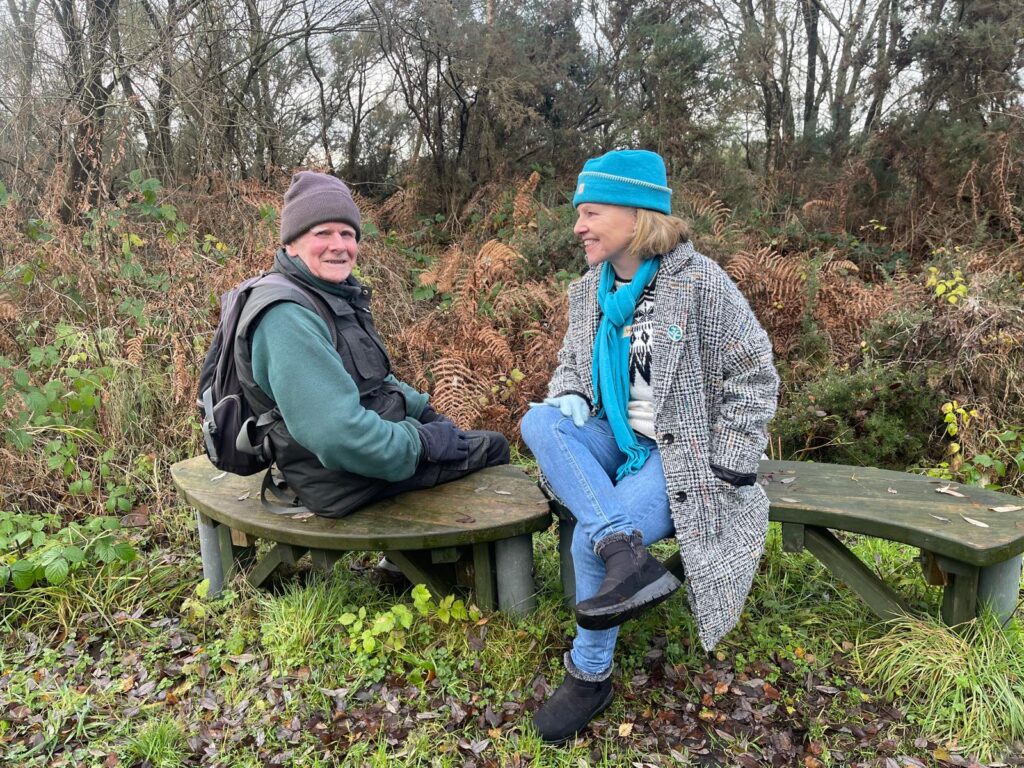 Hugh Shephard and Helen Shaw sitting on wooden benches in front of foliage and trees in the winter.