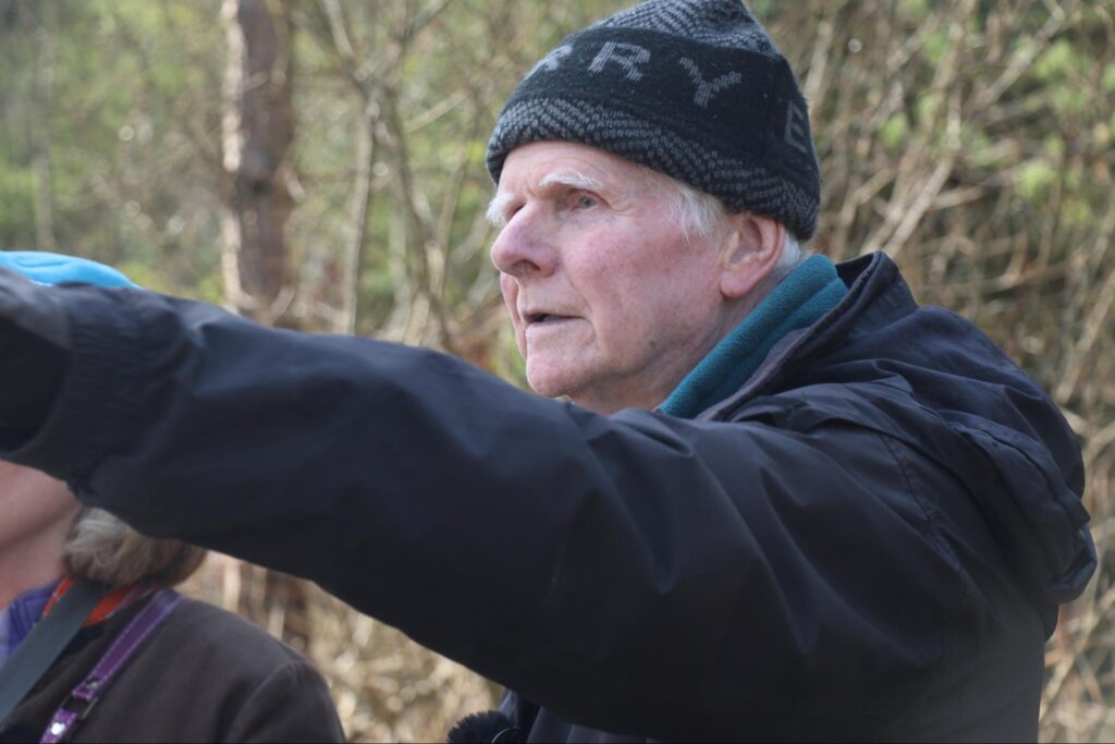 Hugh Shepherd close up photograph dressed in jacket and woolly hat pointing towards something out of frame.