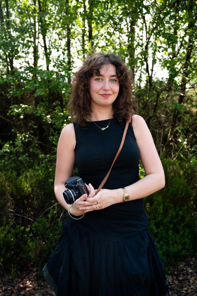 Woman, Evanna Devine, holding a DSLR camera by her side and standing in front of foliage and trees smiling at a camera.