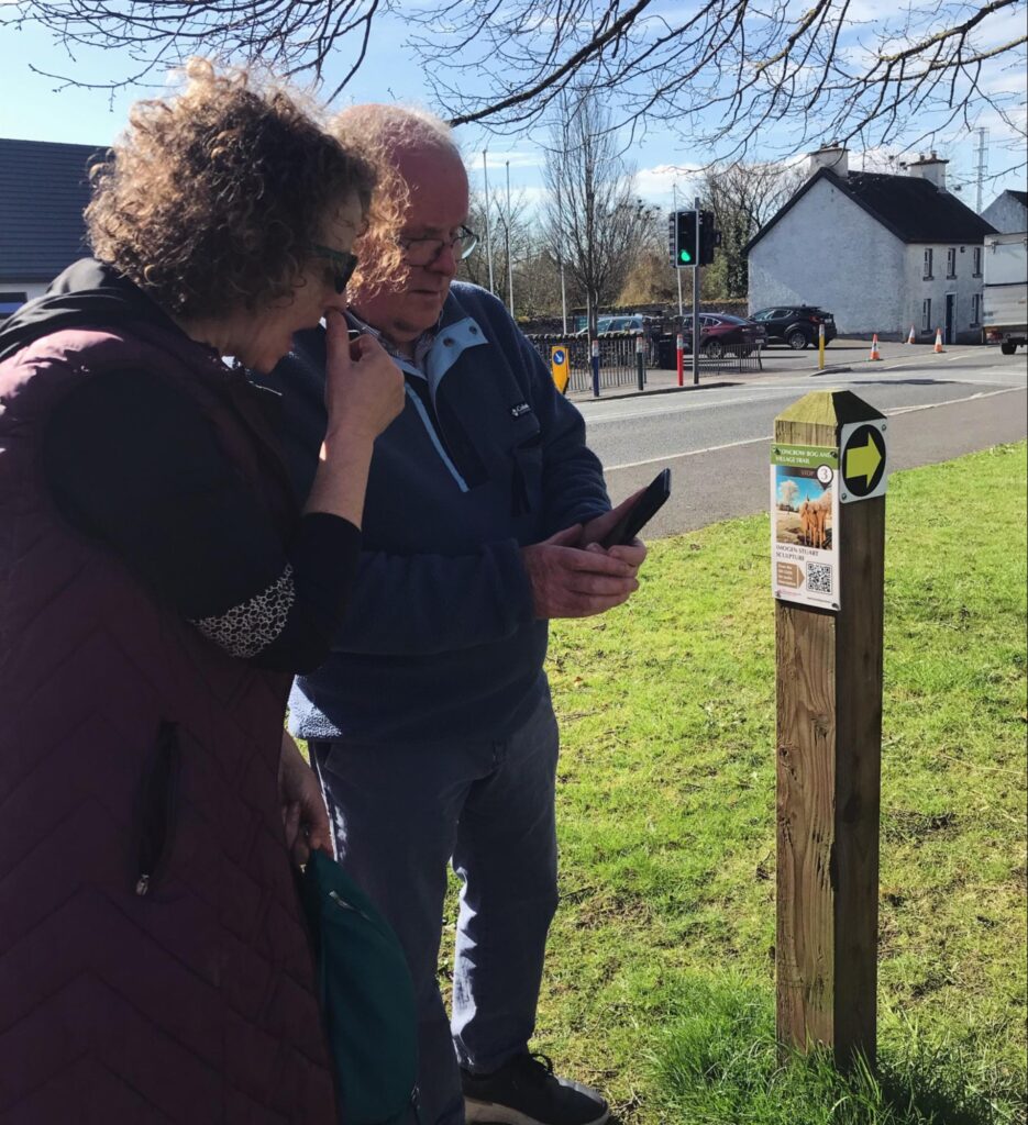 Eugene Dunbar and Annie Holland standing a wooden pole looking at a mobile phone in Eugene's hand. Eugene is explaining how to use QR codes.