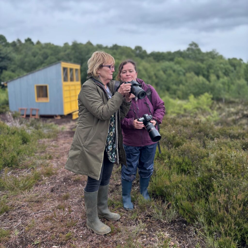 Two women standing on a bog each holding a camera. Both looking at one camera in one of the women's hands looking at photo just taken.