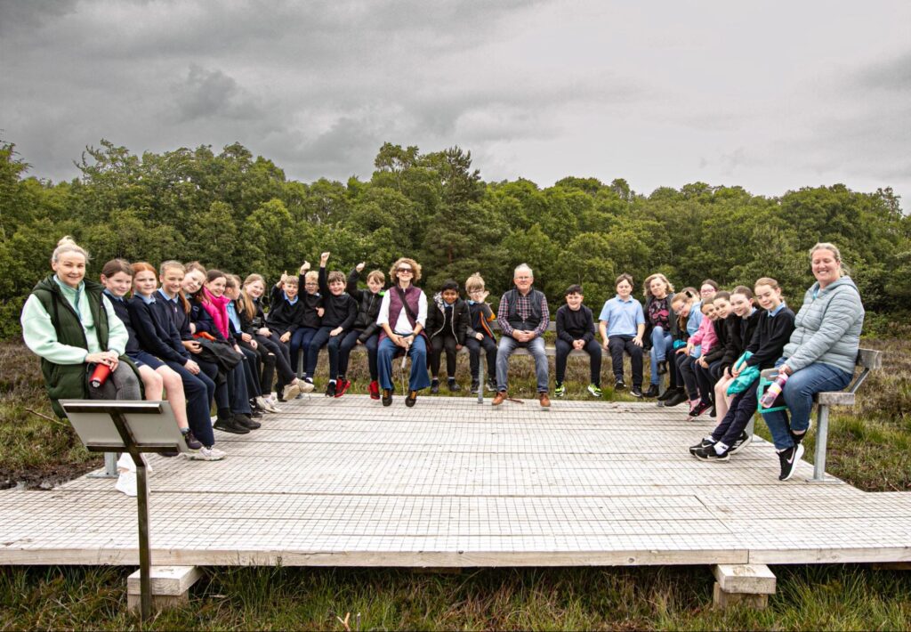 Group of adults and primary school children sitting on a platform on the bog smiling at a camera.