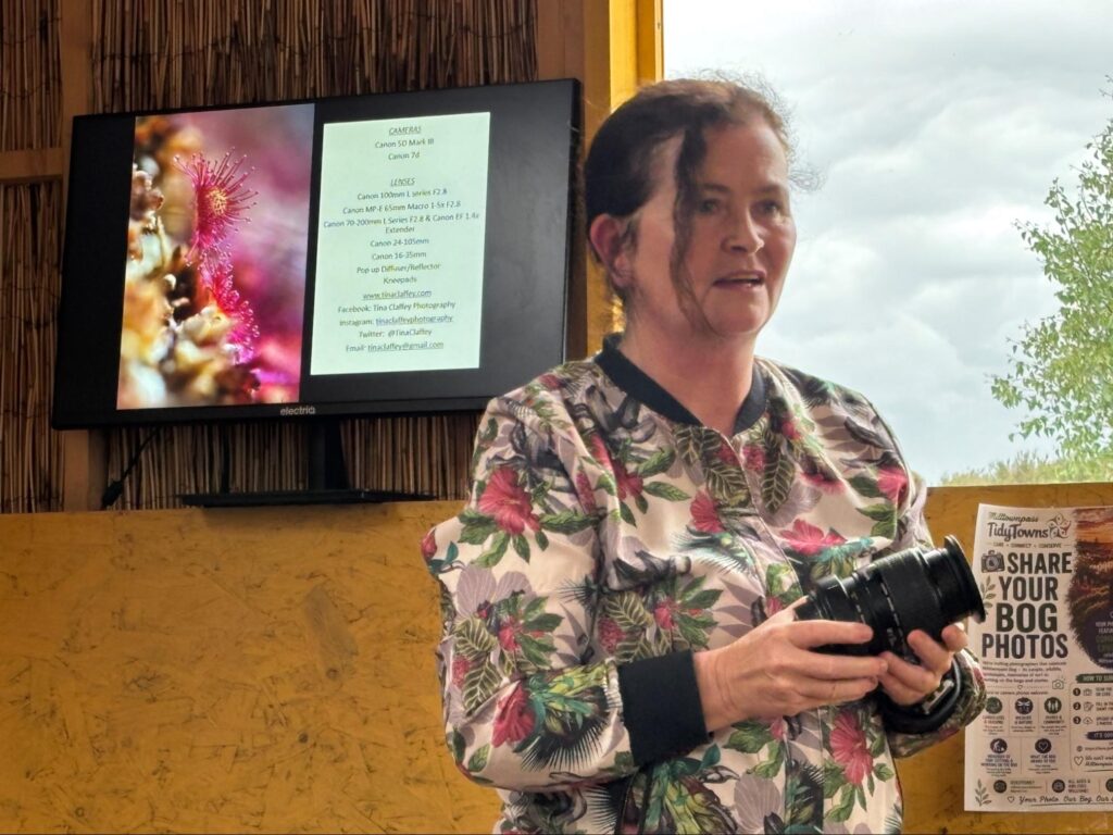 Woman standing in front of a tv showing a presentation. She is holding a camera and speaking to a group outside of frame.