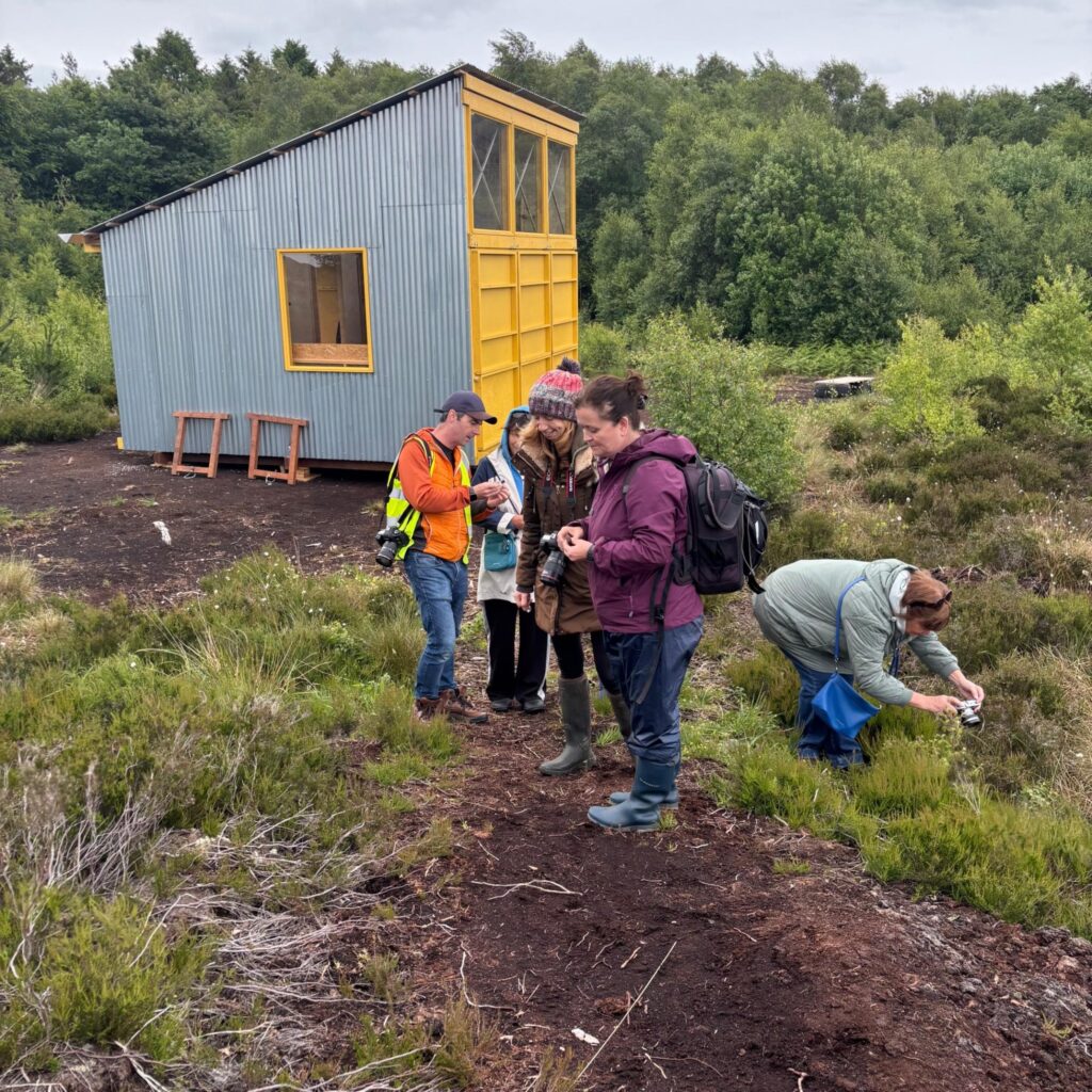 Group of people gathered outside Bog Bothy on a bog holding DSLR cameras and taking photos of plants.