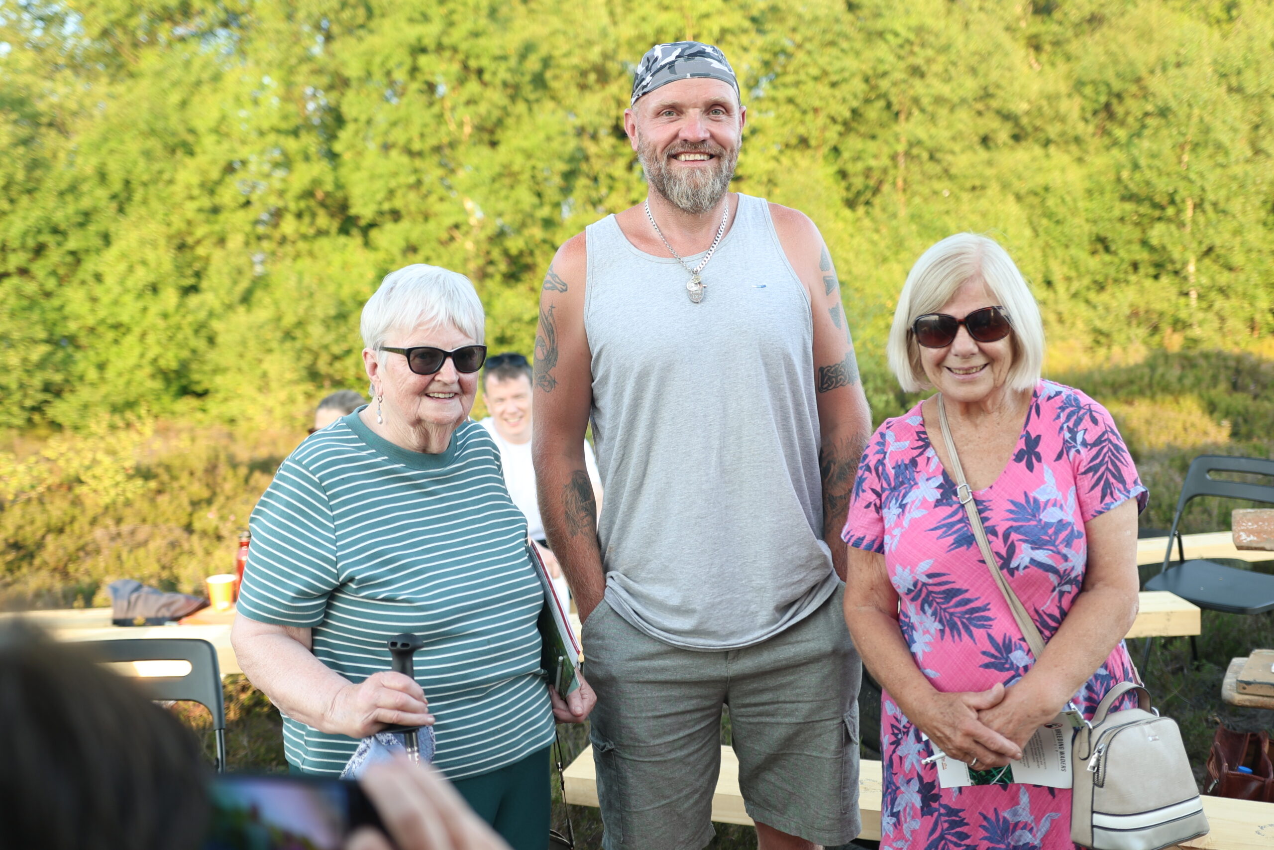 Two women, wearing sunglasses, smiling and standing either side of a taller man. 