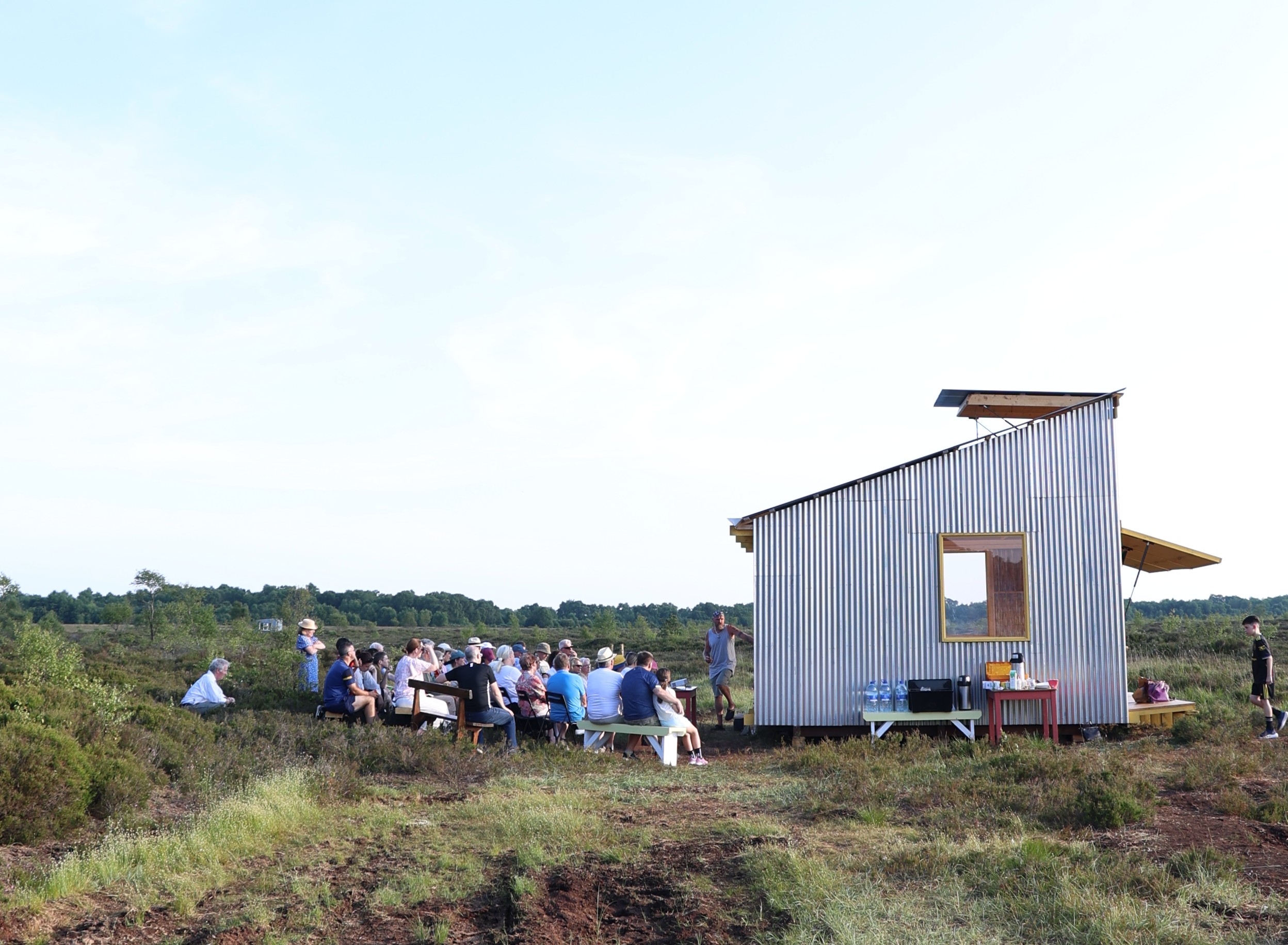 To the left, crowd of people sit on benches facing the Bog Bothy.  