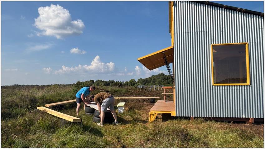 Two men lift tyres and wood, creating a bench in front of the Bog Bothy. 