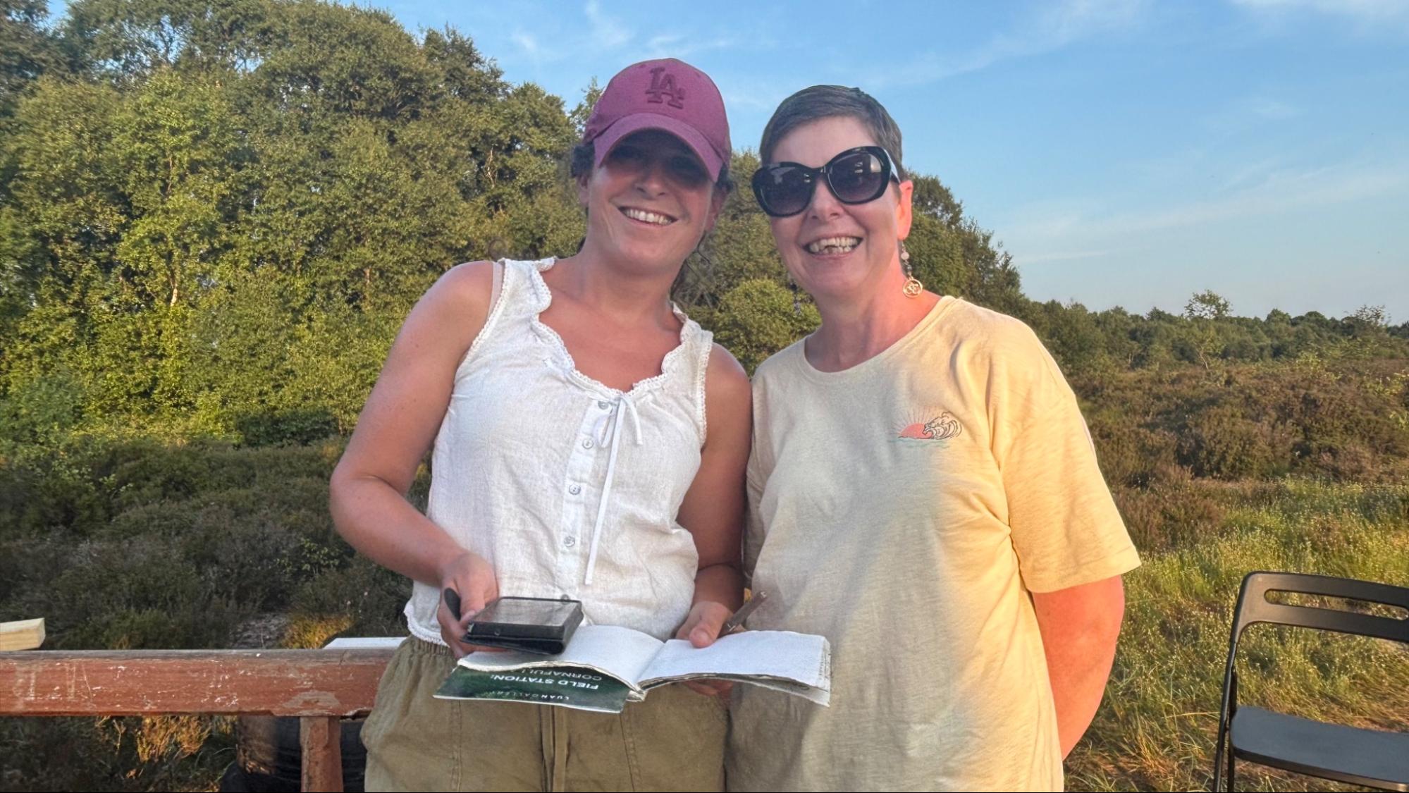 Two women smile while one holds a notebook and a phone in her hand. 