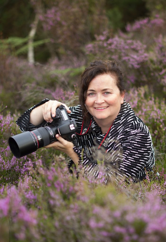 Woman sitting among plants in a bog and holding a camera while smiling at the camera