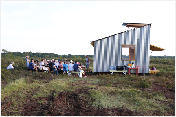 Landscape image of bog bothy with crowd of people sitting outside listening to a speaker