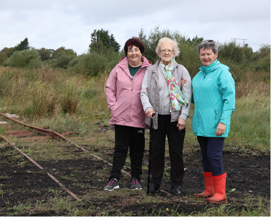 Three women standing looking straight ahead and on an old rail line in a bog.